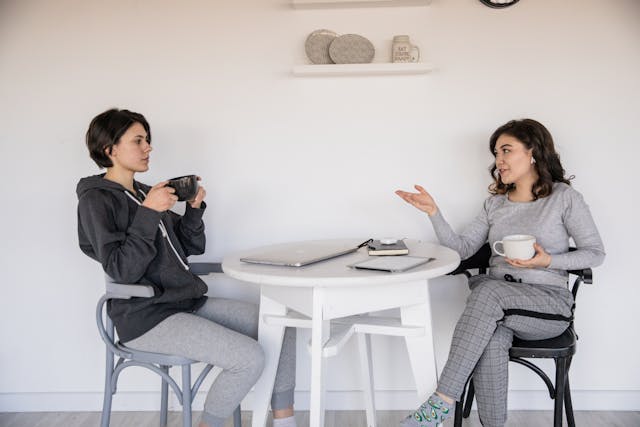 two-people-sitting-at-a-white-table-having-a-conversation-holdig-coffee-cups