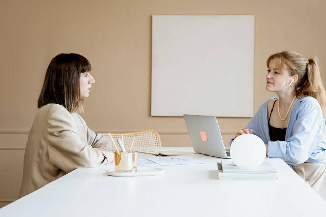 two people sitting at a white table having a work conversation with laptops pens and notebooks