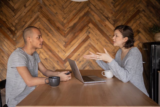 two people sitting at a wooden table with coffee cups and a laptop in front of one person